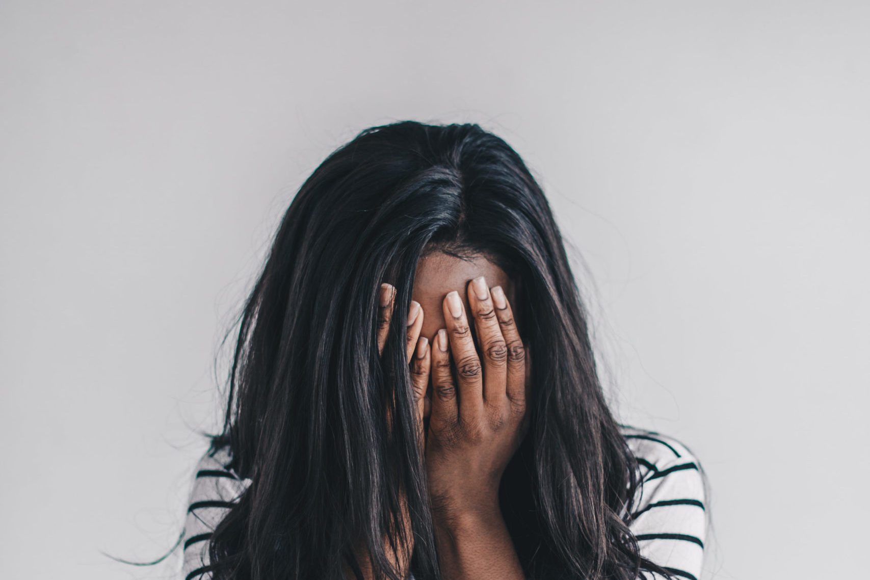 Frau mit lange schwarzen Haaren uns gestreiften Shirt bedeckt ihr Gesicht mit beiden Händen